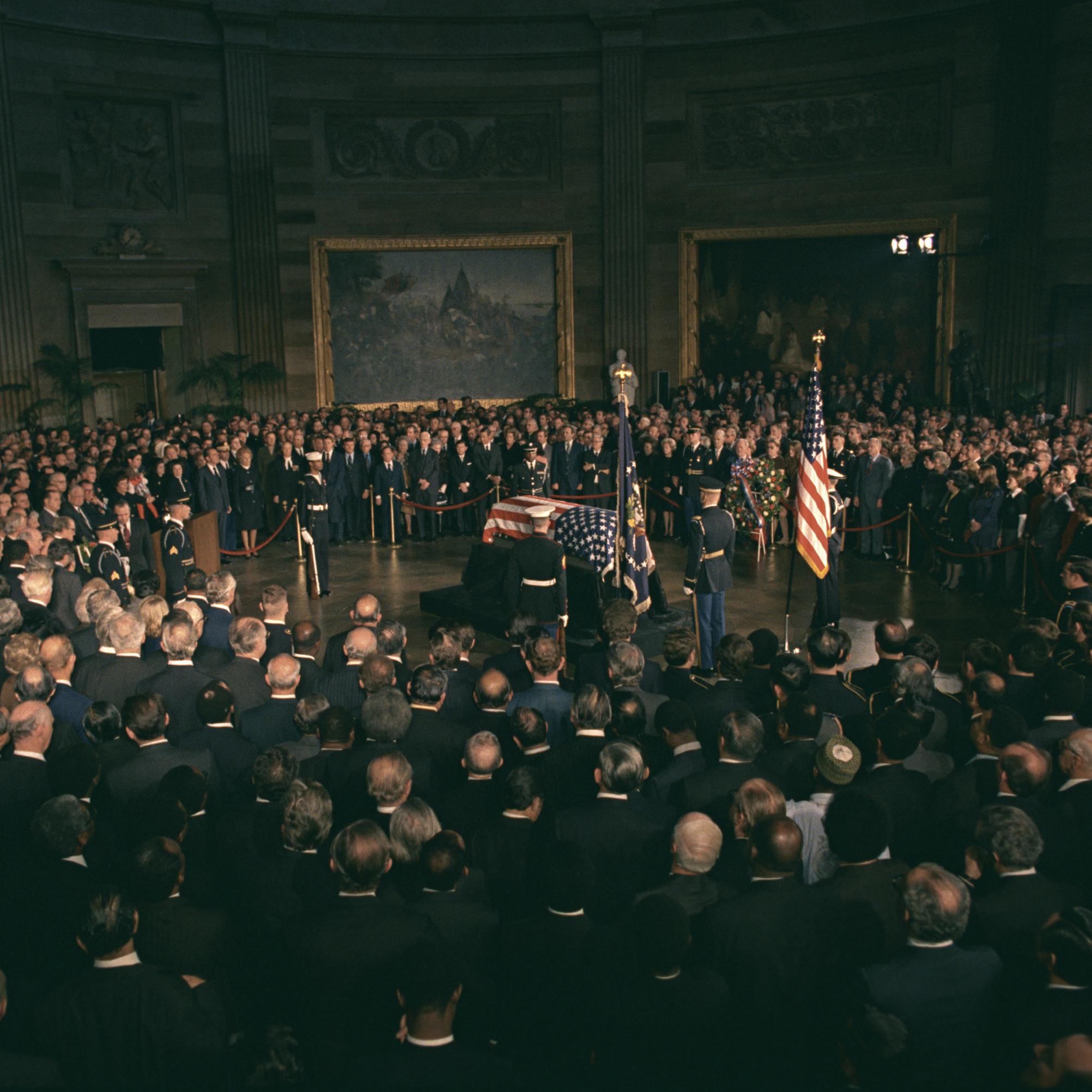 President Richard M. Nixon, members of Congress, and others paying respects to President Lyndon B. Johnson as he lies in state. The casket, draped in an American flag sits in the center of a room, a crowd encircling it.